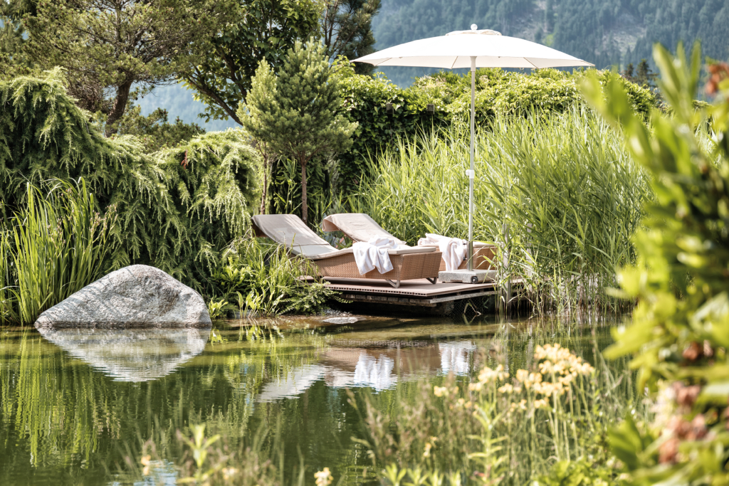 Botanic Garden der Wellnessresidenz Alpenrose mit Teich, üppigem Grün und zwei Liegen mit Sonnenschirm